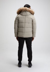 Doudoune beige avec une capuche en fourrure, présentant des sections matelassées. Portée avec un pantalon noir et des chaussures noires, sur un fond uni.