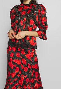 Blumenbluse mit Puffärmeln, schwarzer Stoff mit roten Blumenmustern, Lochstickereien und einem gerafften Saum. Kombiniert mit einem passenden Rock.