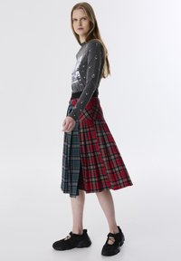 Langer karierter Rock mit roten und grünen Abschnitten, mit Faltenwurf, kombiniert mit einem grauen Langarmshirt, das mit einem Grafikdruck verziert ist. Schwarze Sneakers.