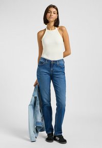 Top halter blanco con textura acanalada, jeans de mezclilla azules y zapatillas negras; sosteniendo una chaqueta de mezclilla azul clara. Diseño simple y ajustado con un aspecto clásico.