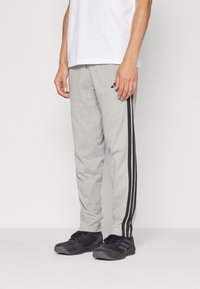 Grijze sportbroek met zwarte zijstrepen, gemaakt van een zachte stof, gecombineerd met zwarte schoenen. Model draagt een eenvoudig wit t-shirt.