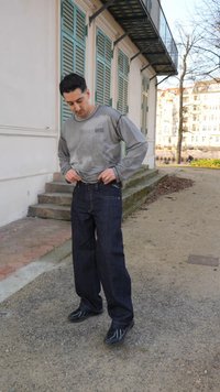 Mann in grauem Langarmshirt und dunkler Jeans, der draußen bei Sonnenschein nahe Treppen und verschlossenen Fenstern seine Hose richtet.
