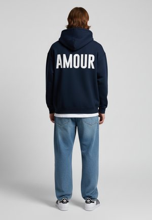 Sweat à capuche marine avec le texte blanc "AMOUR" dans le dos, porté sur une chemise blanche. Associé à un pantalon en denim bleu clair et des baskets noires.