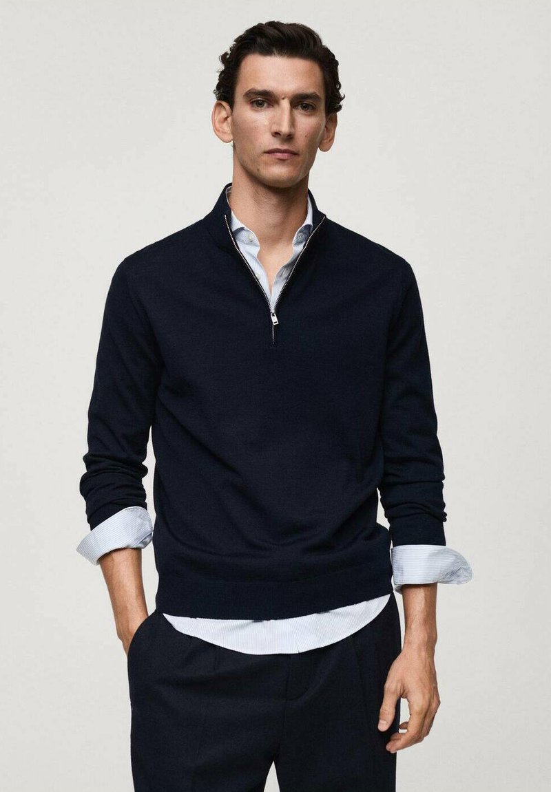 Navy blue zip-neck sweater made of a soft knit fabric, featuring a ribbed hem and cuffs, layered over a light blue collared shirt.