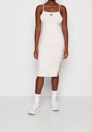 Femme portant une robe midi blanche ajustée avec de fines bretelles, un petit logo sur la poitrine, des chaussettes blanches Nike et des baskets blanches.