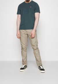 Mørk teal bomulds T-shirt med korte ærmer og et lille rødt logo, parret med beige bukser og sorte og hvide lavt top sneakers.