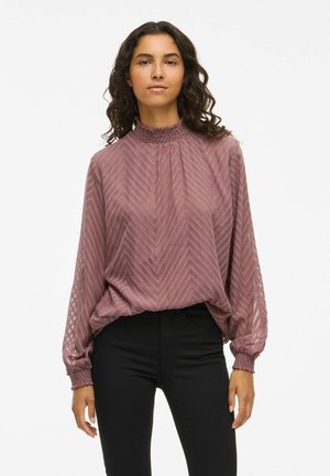 Blouse à manches longues, d'un rose doux, avec un motif chevron texturé, un col smocké haut et des poignets élastiques, associée à un pantalon noir ajusté.