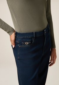 Pantalones de mezclilla oscura con un diseño ajustado, que cuentan con un botón decorativo en el bolsillo y una textura suave. Acompañados de una blusa verde oliva.