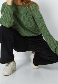 Grüner Strickpullover mit strukturiertem Muster, Rundhalsausschnitt und lockeren Ärmeln, kombiniert mit schwarzen Weitbein-Hosen und hellen Sneakern.