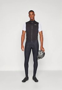 Black cycling vest with a zip closure, paired with a short-sleeve white top and black leggings. Holding a black helmet, wearing cycling shoes.