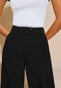 Pantaloni neri su misura a vita alta realizzati in tessuto liscio, con chiusura frontale a bottone, pinces e tasche.