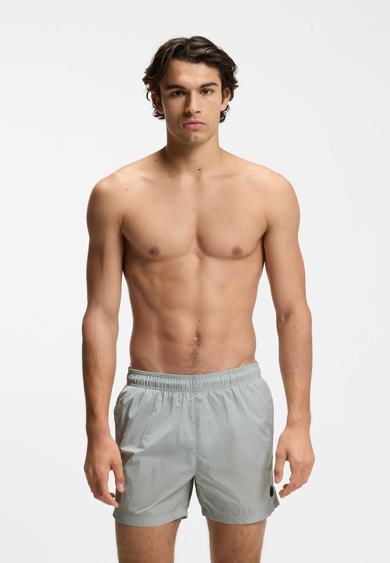 Jeune homme aux cheveux courts et foncés portant un short de bain gris clair, debout sur un fond blanc uni, torse nu et face à l'avant.