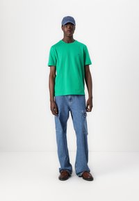 Zelené bavlnené tričko s krátkymi rukávmi, modré džinsové nohavice s vreckami a hnedé topánky. Námořní čiapka dotvára celý outfit.