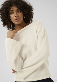 Pull couleur crème à texture douce avec des manches larges et longues et un décolleté bateau, présentant une coupe décontractée et des détails de design minimalistes.