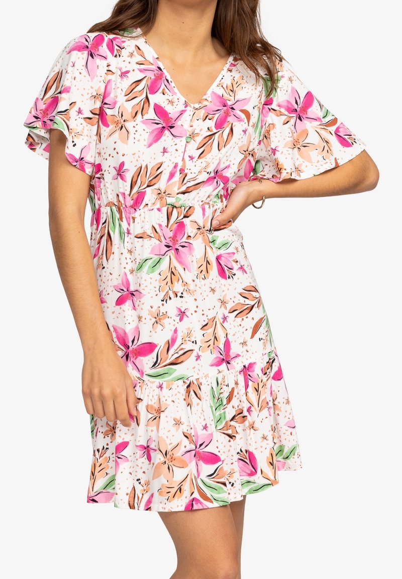 Femme portant une robe blanche à manches courtes avec des motifs floraux roses et pêches, debout avec une main sur la hanche et l'autre détendue.