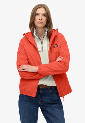 Veste imperméable rouge avec capuche, poches zippées et logo brodé. Portée sur un pull léger gris et un jean en denim bleu.