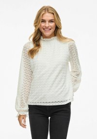 Blouse blanche texturée avec un col montant, des manches longues en tissu transparent et des poignet froncés, présentant un motif en zigzag et un ourlet festonné.