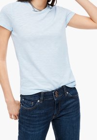 Vaaleansininen lyhythihainen t-paita, jossa on pehmeä tekstuuri, yhdistettynä tummansinisiin farkkuihin, joissa on koristeelliset tuplapainikkeet ja ompeleet.