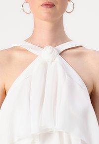 Balts halter-top ar plisēta dizainu un rozes akcentu ap kaklu, izgatavots no mīksta, plūstoša auduma. Gluda tekstūra un minimāls noformējums.