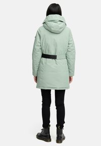 Manteau doudoune à capuche vert menthe avec une taille ceinturée, présentant une texture lisse et des accents de logo sur le col. Porté sur un pantalon noir et des bottes.
