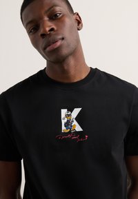 Fekete pamut póló, amelyen a "K" betű grafikai terve és egy hímzett Donald Kacsa látható, alatta pedig egy piros aláírással.
