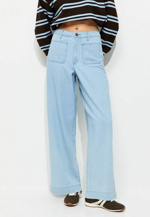 Personne portant un jean large taille haute de couleur bleu clair, un haut à manches longues rayé marron et bleu, coupé court, et des baskets marron avec des semelles blanches.