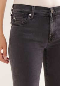 Close-up de una persona que lleva pu jeans gris oscuro de cintura alta con detalles de botones y bolsillos, de pie contra un fondo claro.