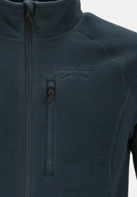 Fleecejakke i m�ørkeblå med høj krave, lynlås foran og en brystlomme med lynlås. Blød tekstur, minimal branding synlig.