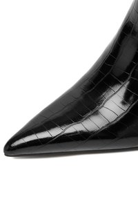 Pantofii cu vârful ascutit din piele patenta neagră, cu un detaliu lucios de textură de crocodil, pe un fundal alb.
