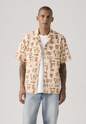 Man wearing short-sleeve beige shirt with western-themed prints over white t-shirt and light blue jeans, standing against plain background.