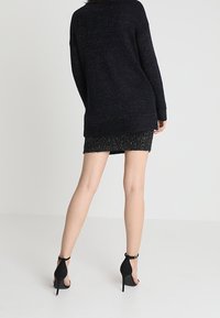 Černý svetr s volnějším střihem, texturovaným úpletem a dlouhou siluetou, zkombinovaný s černou minisukní a podpatky, které zdůrazňují délku nohou.