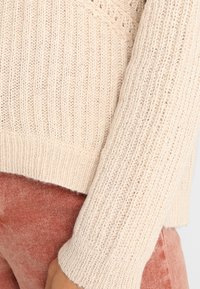 Pletený béžový svetr s jemně vroubkovanou strukturou, který má široký dekolt a dlouhé rukávy, v kombinaci s měkkými růžovými kordovými kalhotami.