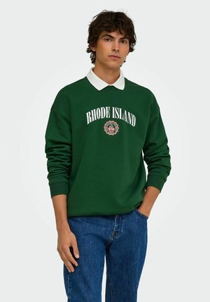Jeune homme aux cheveux bouclés portant un sweat-shirt Rhode Island vert foncé par-dessus une chemise blanche à col et un jean bleu, debout avec une main dans la poche.