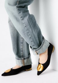 Ballerine in camoscio nero con punta affilata, caratterizzate da un grande accento circolare dorato e dettaglio di perline dorate sul tallone. Indossate con jeans di denim risvoltati.