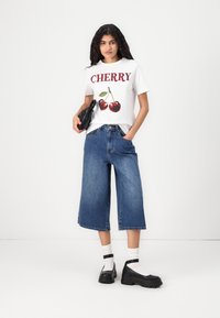 Balta T-krekls ar sarkano tekstu "CHERRY" un ķirša grafiku, savienots ar brīviem ziliem denima culottes un melnajām platformas kurpēm, turēdams melnu somu.