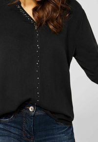 Schwarzes Shirt mit V-Ausschnitt, versehen mit einer Reihe silberner Nieten entlang der Knopfleiste. Kombiniert mit blauen Jeans. Weicher Stoff.