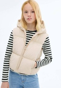 Beige Steppweste mit Kapuze, Reißverschluss und gestepptem Muster; getragen über einem schwarz-weißen gestreiften Langarmoberteil. Lässige Denim-Hosen.