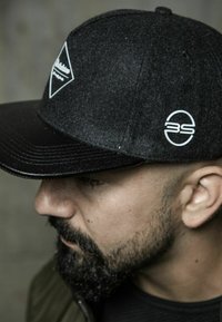 Black felt cap with a wide glossy brim, featuring a white geometric logo and circular emblem, set against a neutral background.