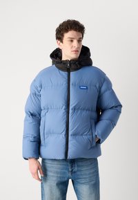 Padded jacket in light blue with a black hood and zip closure. Features two front pockets and a blue logo patch on the chest.