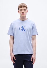 Jasnoniebieski bawełniany t-shirt z dużym niebieskim logo "CK" na środku oraz napisem "Calvin Klein" poniżej. Klasyczny okrągły dekolt i krótkie rękawy.