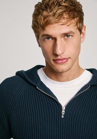 Tamsiai mėlynas megztinis su užtrauktuku priekyje ir gobtuvu. Yra raukšlėta tekstūra ir kontrastinga balta marškinėlė po juo.