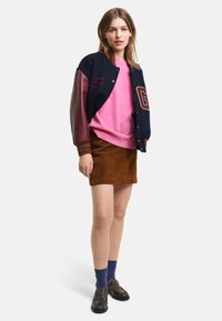 Marineblau und burgunderfarbener Bomberjacke mit Buchstabendetails, rosa Sweatshirt, brauner Wildlederrock, blaue Socken und schwarze Schuhe.