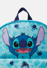 Sac à dos bleu présentant un personnage de dessin animé avec de grandes oreilles et des yeux. Le matériau est lisse, avec un arrière-plan à motifs en bleu clair et des accents plus foncés.