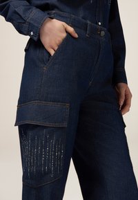 Pantaloni in denim blu scuro con tasca laterale, cuciture a contrasto e accenti decorativi di perline argentate lungo la gamba.