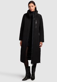 Black long coat with a hood, featuring a front zipper, side pockets, and a sleek design. Worn over a black outfit and knee-high boots.