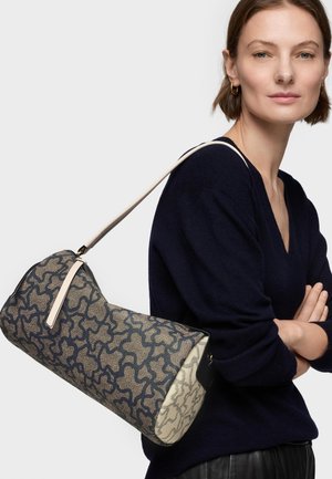 Mujer con cabello corto y castaño, vestida con un suéter azul marino oscuro, llevando un bolso cilíndrico estampado con una correa clara sobre el hombro.