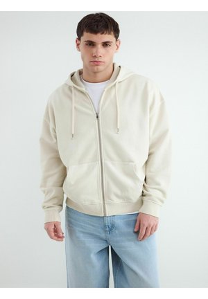 Młody mężczyzna w jasnobeżowej bluzie na suwak nałożonej na białą koszulę i jasnoniebieskie dżinsy, stojący na jednolitym białym tle.