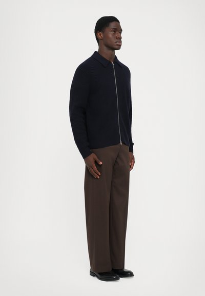 Hombre de pie con un suéter azul marino con cremallera, pantalones de pierna ancha marrones y zapatos negros, delante de un fondo blanco liso.