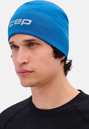 Giovane uomo che indossa un berretto aderente di colore blu brillante con il logo bianco "cep", abbinato a una maglietta nera a girocollo, che guarda leggermente a sinistra.