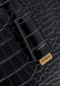 Cuir noir texturé crocodile avec une sangle polie et une plaque métallique dorée gravée de « GUESS » sur la surface d’un sac à main.